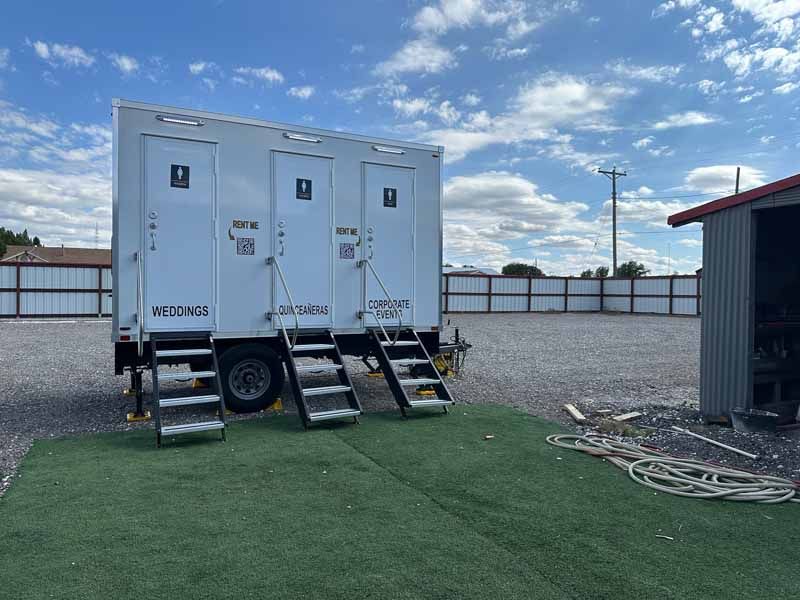 A white portable restroom trailer with three individual stalls and steps, parked on gravel next to a small building.