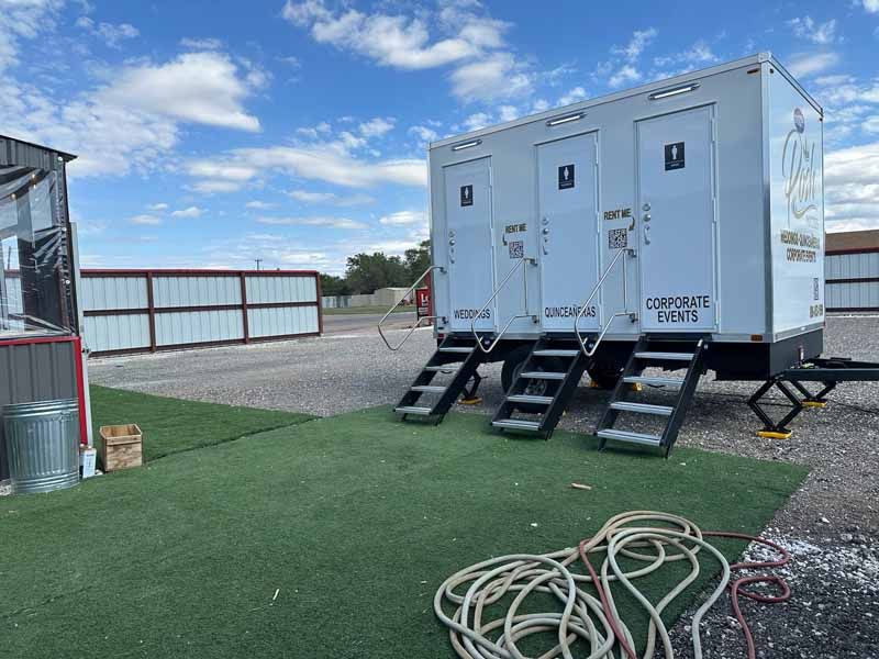 White portable restroom trailer with three doors and steps on green turf.