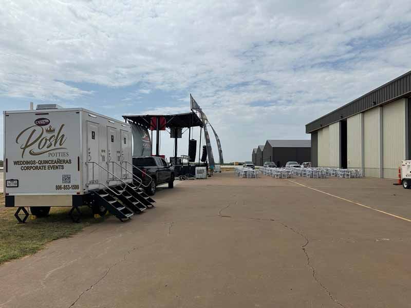 Mobile restroom trailer, stage, and chairs set up on an airfield under a cloudy sky.