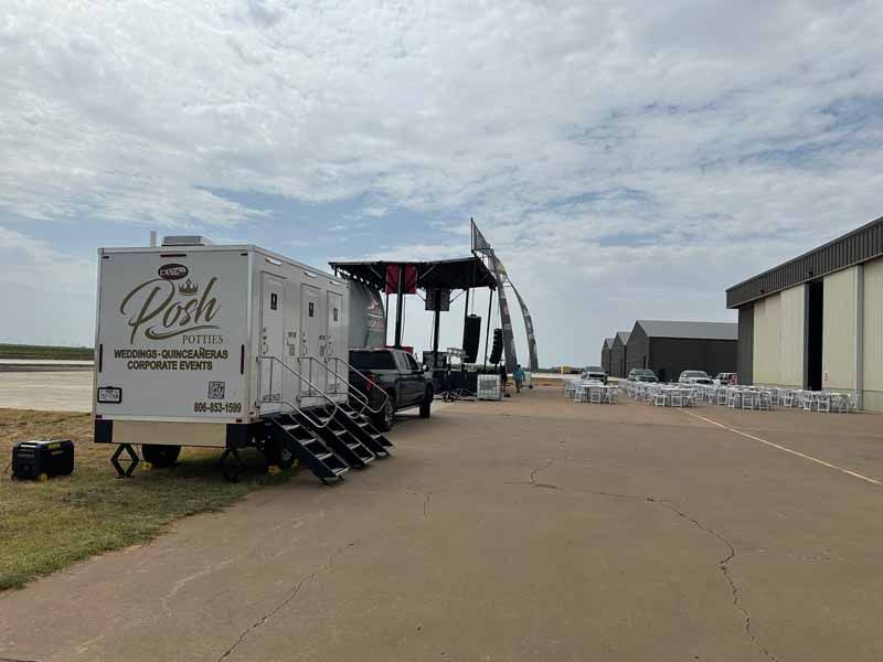 Portable restrooms with a stage on an airfield with seating and a building visible.
