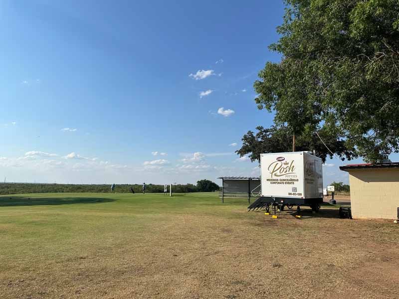 Sunny outdoor scene with a white trailer, trees, and green grass. Blue sky with a few clouds in the background.