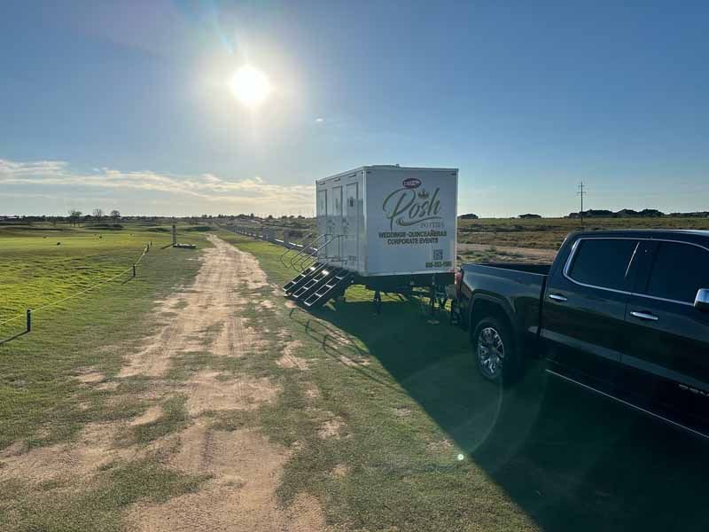 A portable restroom trailer with a black truck parked next to it on a dirt road by a golf course.