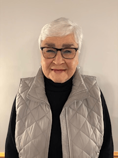An elderly woman wearing glasses and a vest is smiling for the camera.