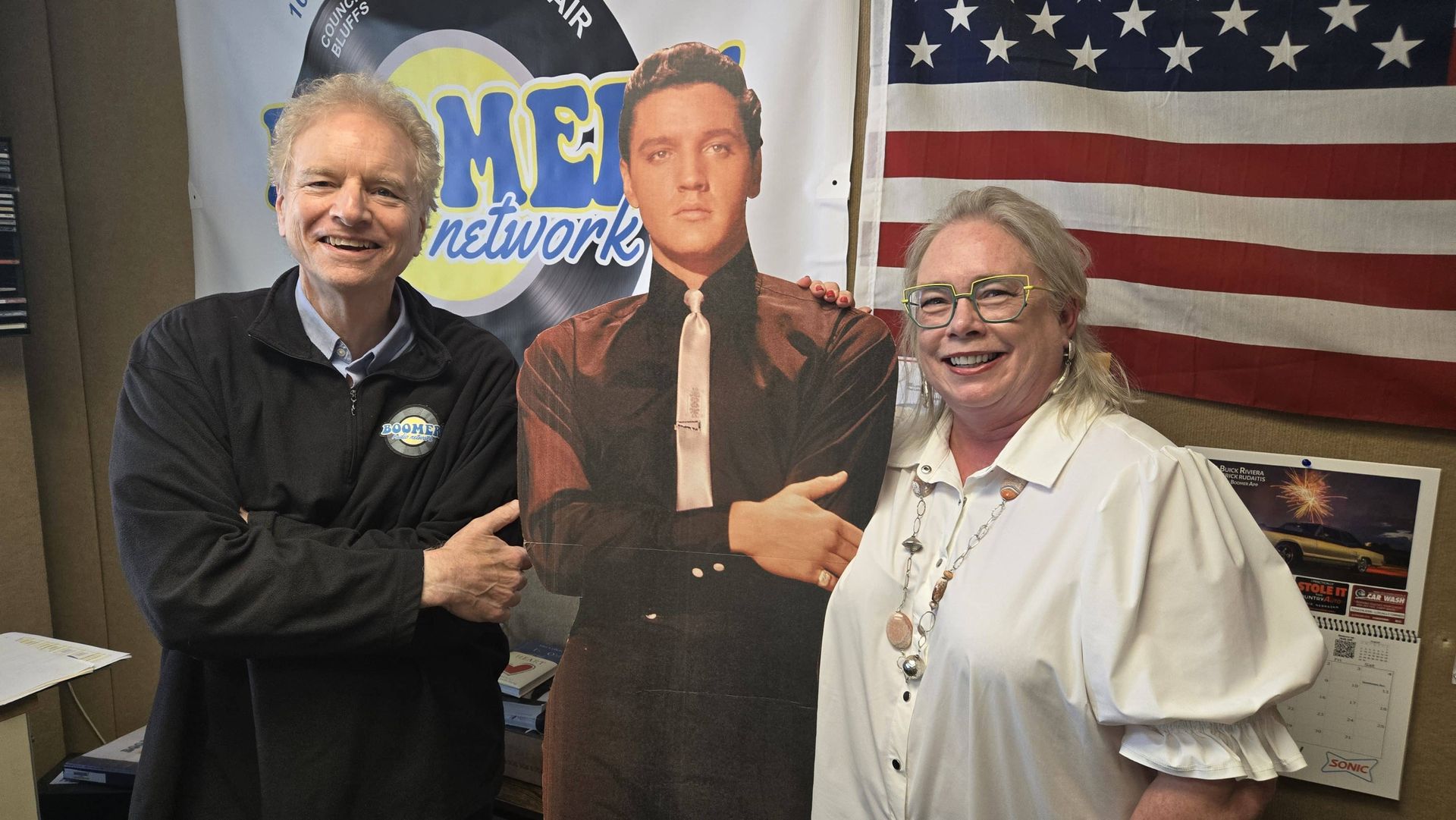 Two people posing with a cardboard cutout in front of a U.S. flag and radio station sign