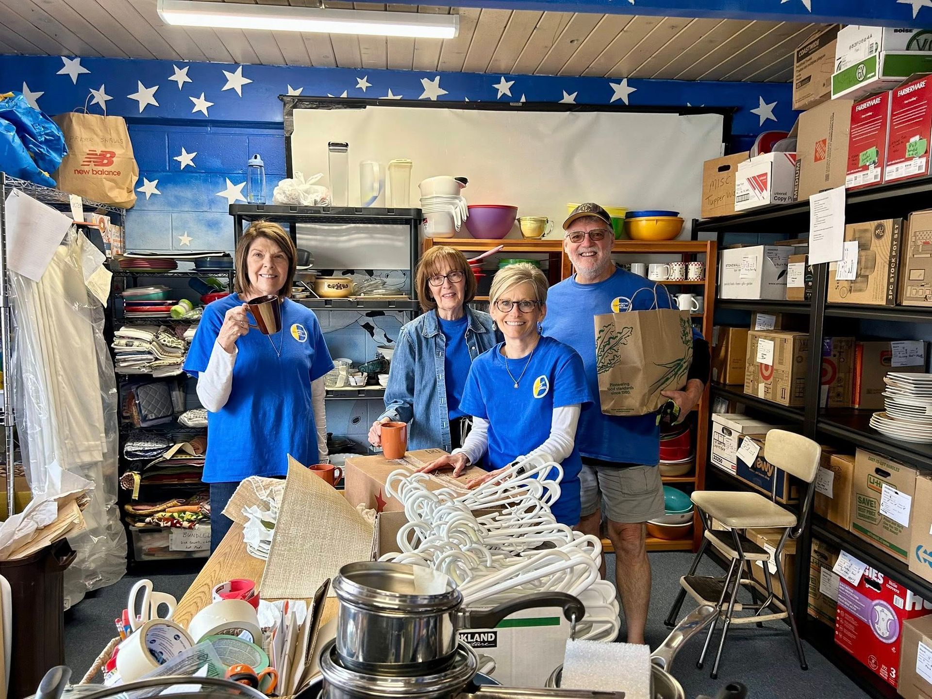 Four people in blue shirts sorting through donations in a cluttered room with shelves and a star-patterned wall.