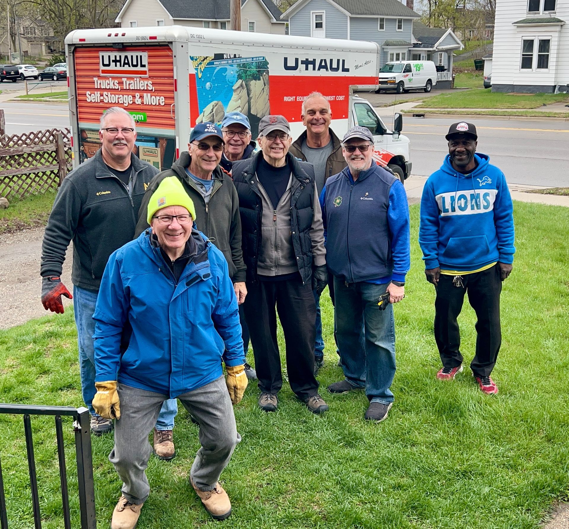 Group of men standing in front of a U-Haul truck and a house, smiling. Green grass and overcast sky.