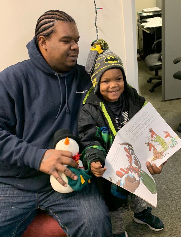 Father and child reading a book. The child smiles, wearing a hat and jacket. A snowman toy sits nearby.
