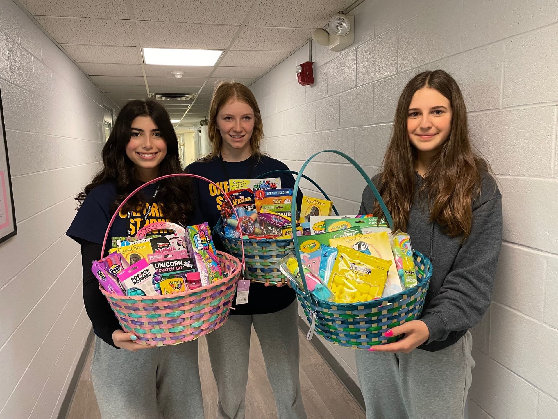 Three people hold colorful gift baskets in a hallway. They are smiling.