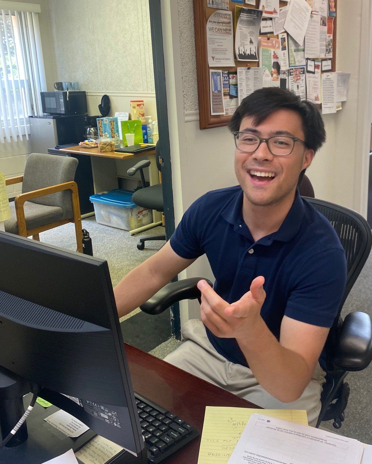 Man smiling and gesturing near a computer in an office setting.