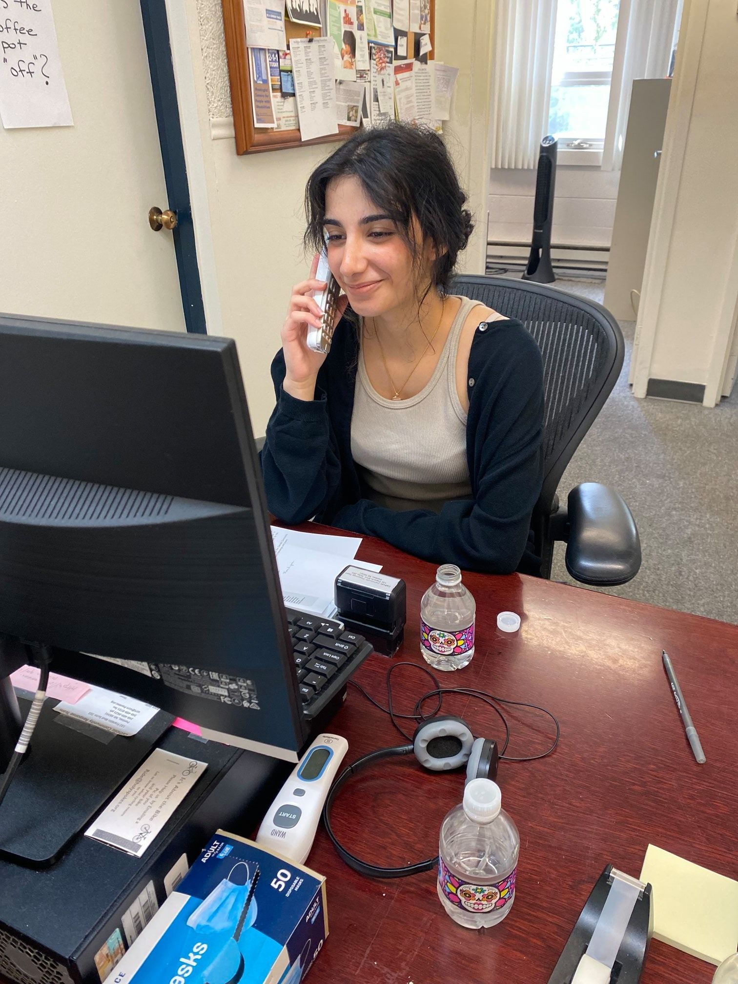 Woman on a phone, sitting at a desk with a computer and water bottles. She's smiling in an office.