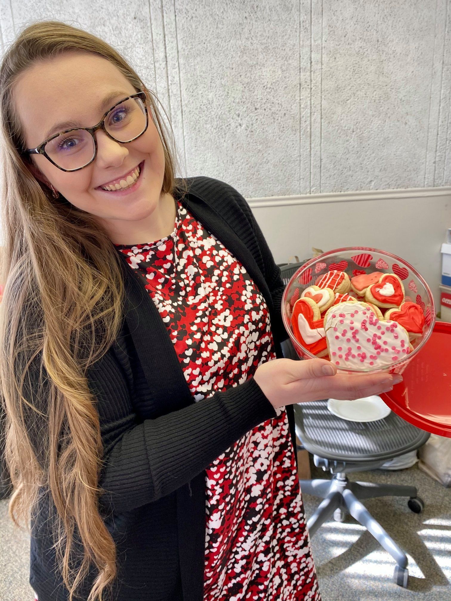 Woman smiles, holding a clear container of heart-shaped cookies. Red and white patterned dress, black cardigan.