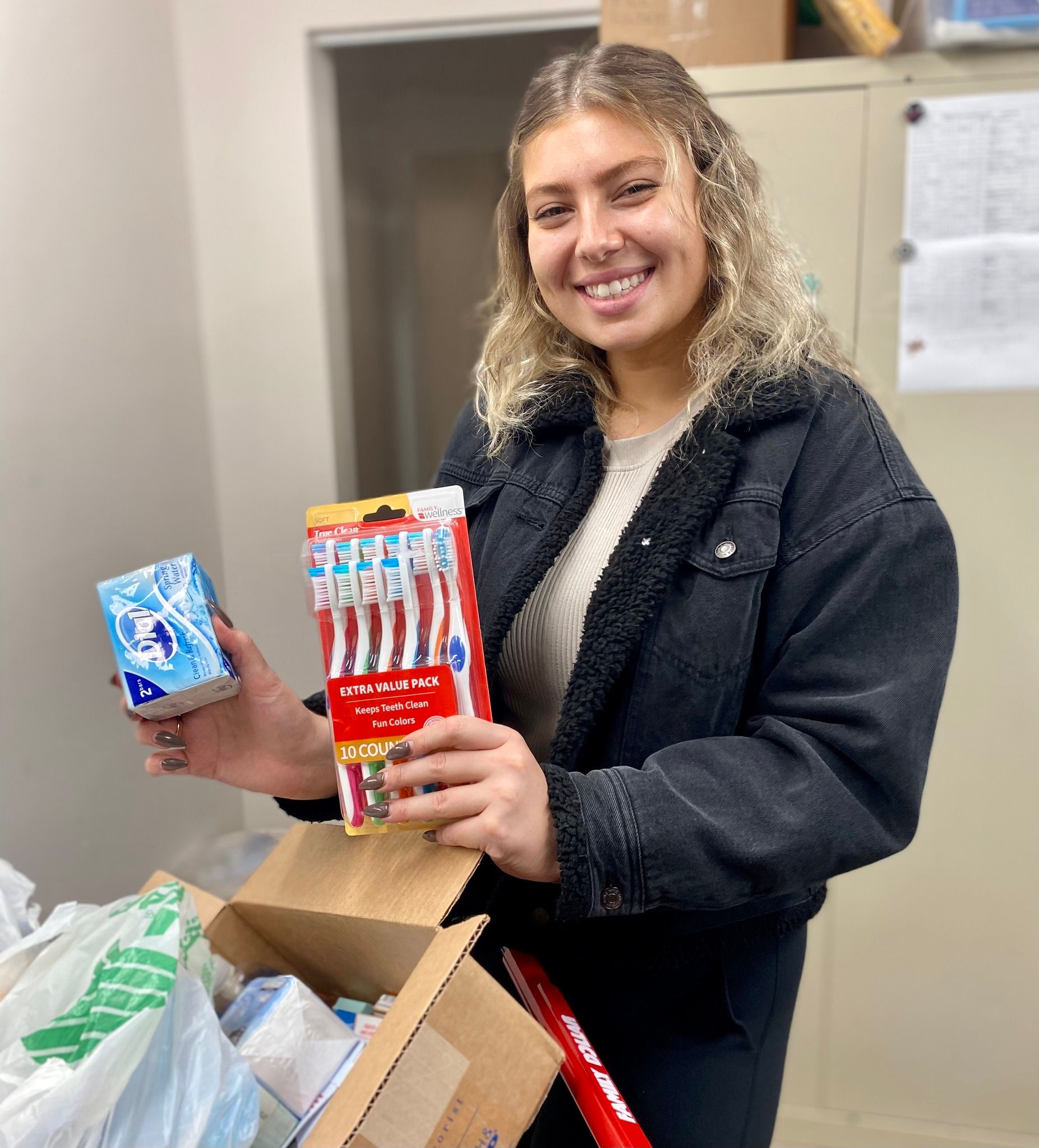 Woman holding toothpaste and toothbrushes, smiling, near open box of items.