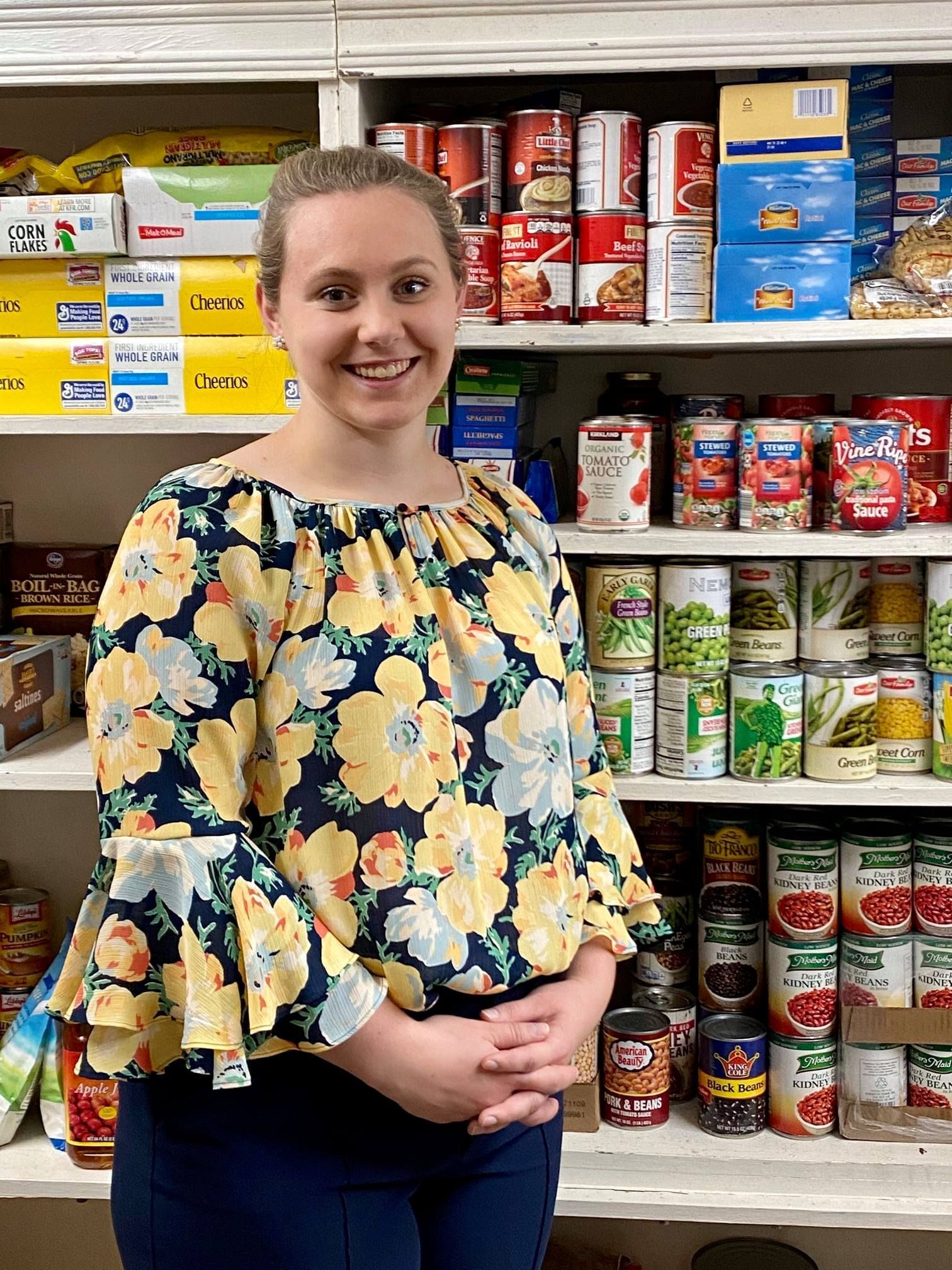 Woman smiles, stands in front of pantry shelves filled with canned goods. She wears a floral shirt, arms crossed.