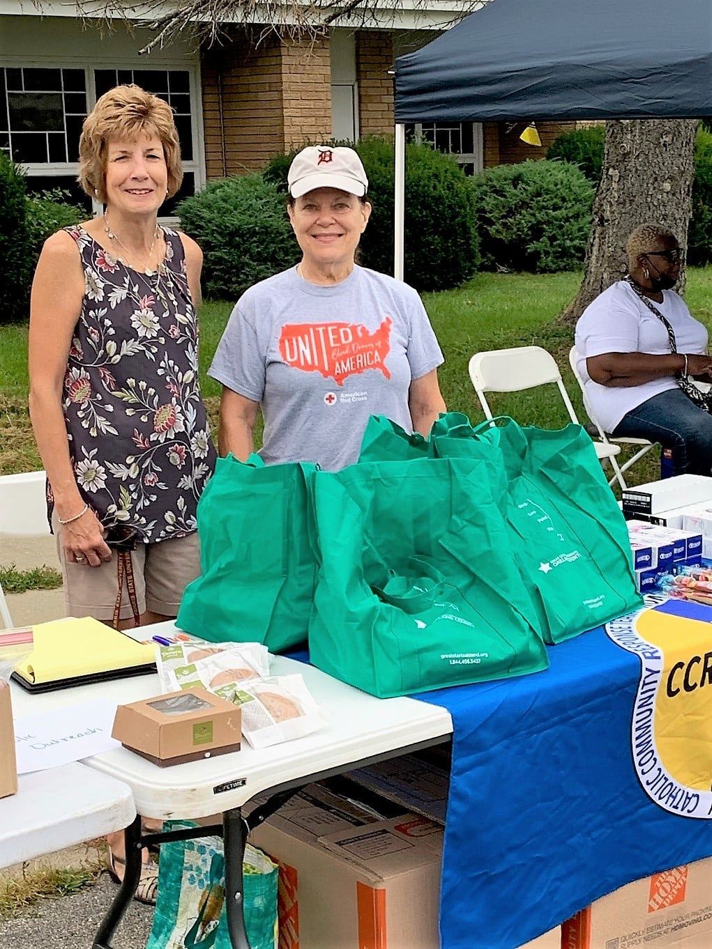 Two women stand behind a table with green bags and pamphlets; outdoor event.