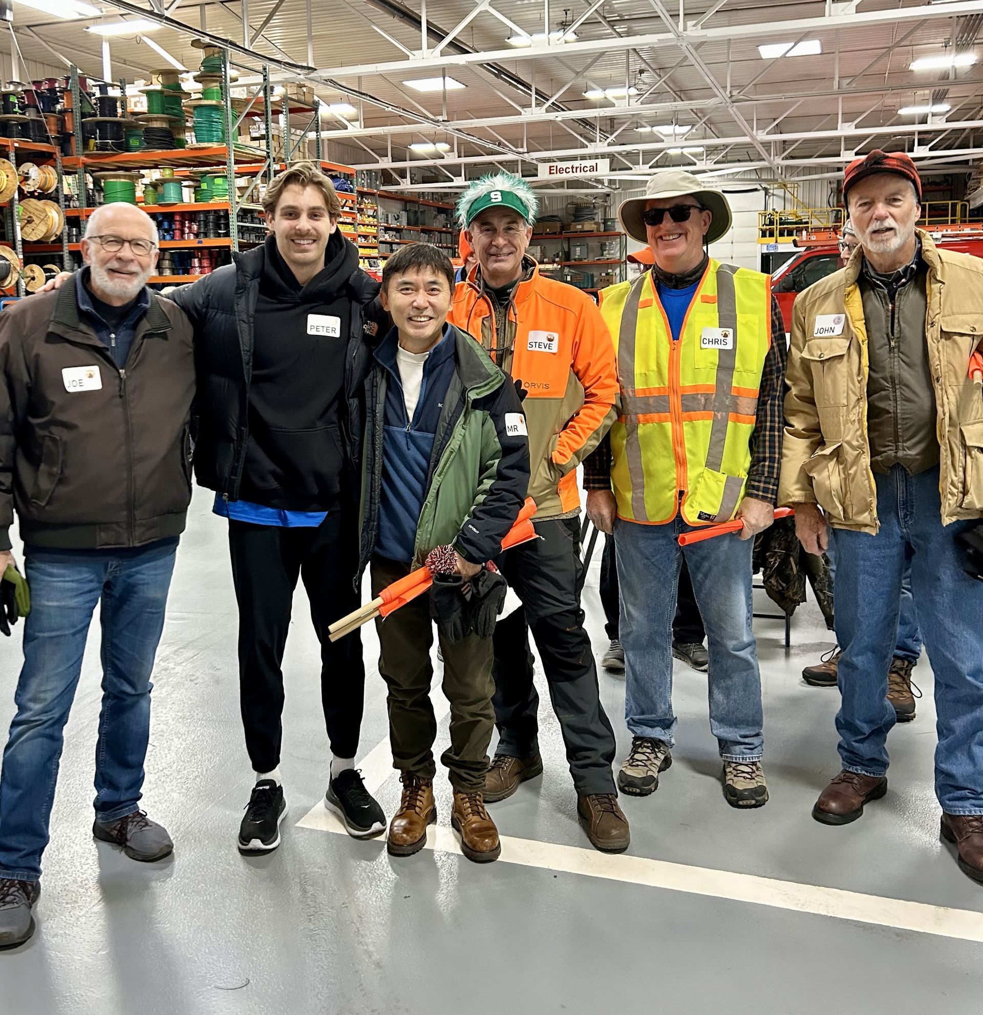 Group of people smiling in a warehouse, some in safety vests, holding orange wands.