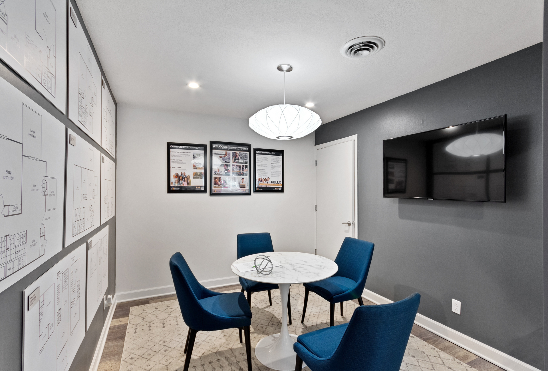 Small communal lounge with a round marble table, blue chairs, wall TV, and framed diagrams along the left wall.