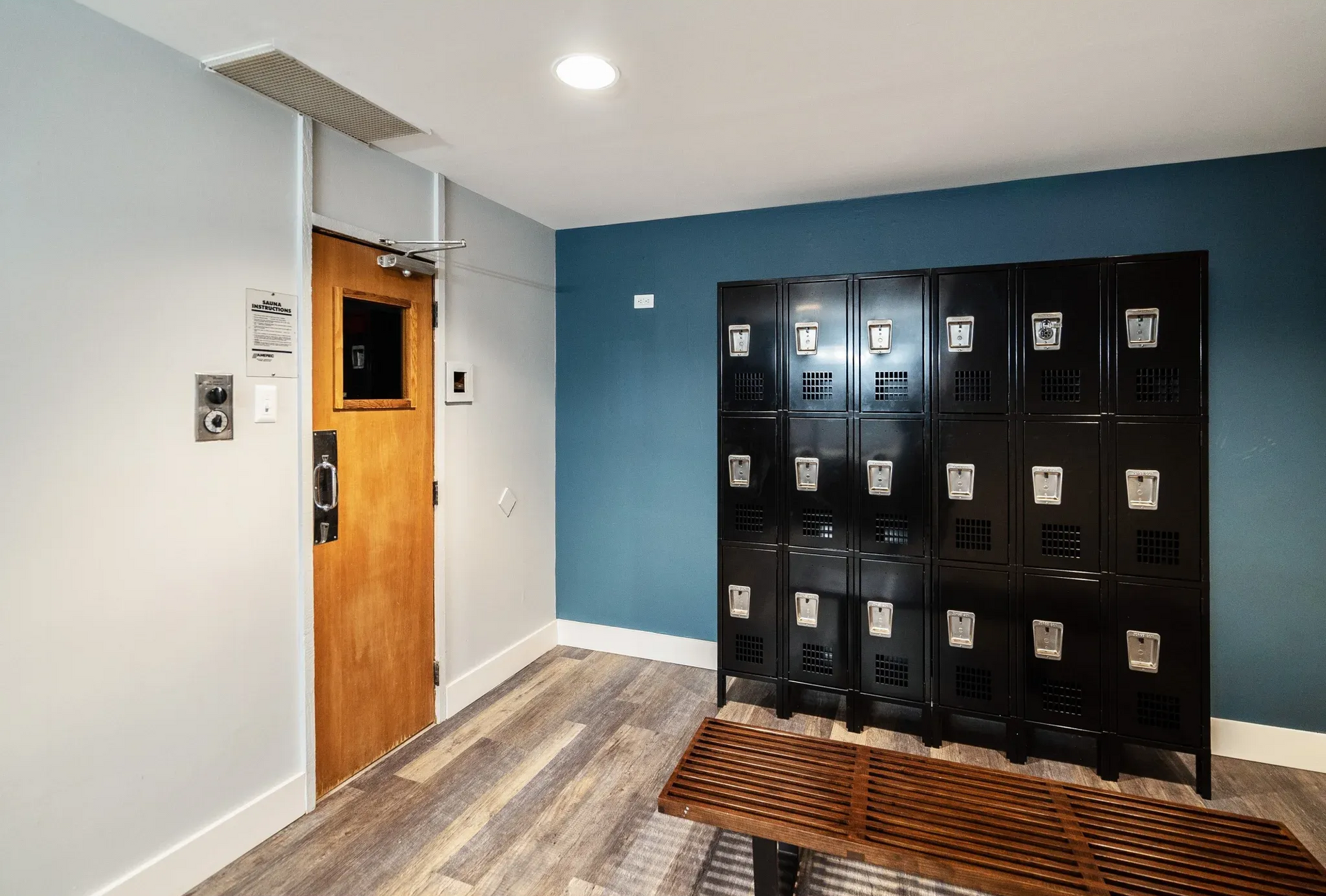 Interior corridor with blue wall, a row of black lockers, and a wooden bench.