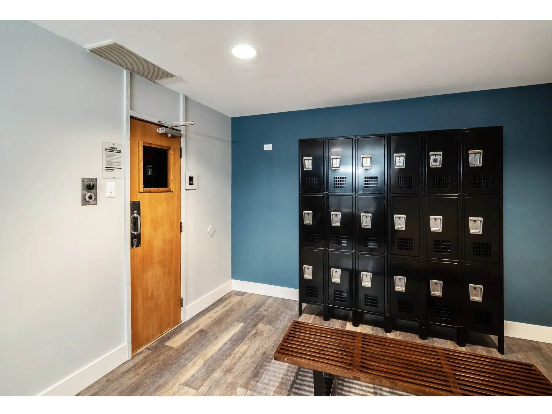 Interior corridor with blue wall, a row of black lockers, and a wooden bench.