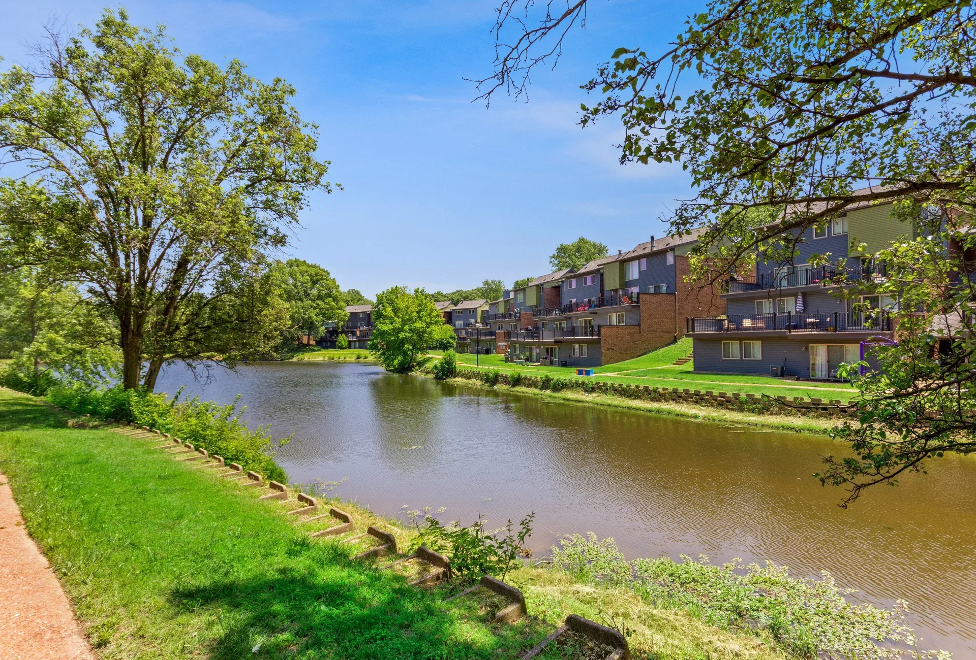 Exterior view of apartment buildings along a river with trees and a green lawn.