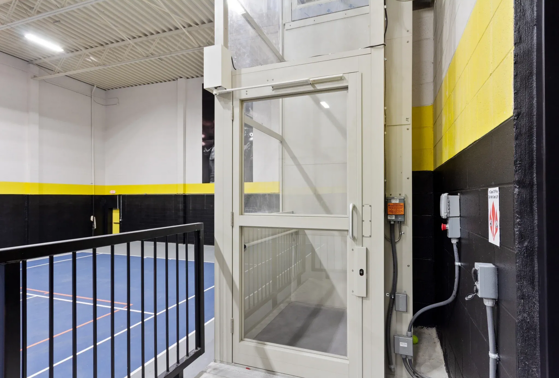 Indoor community recreation area with a blue court floor, yellow/black walls, and a glass service elevator.