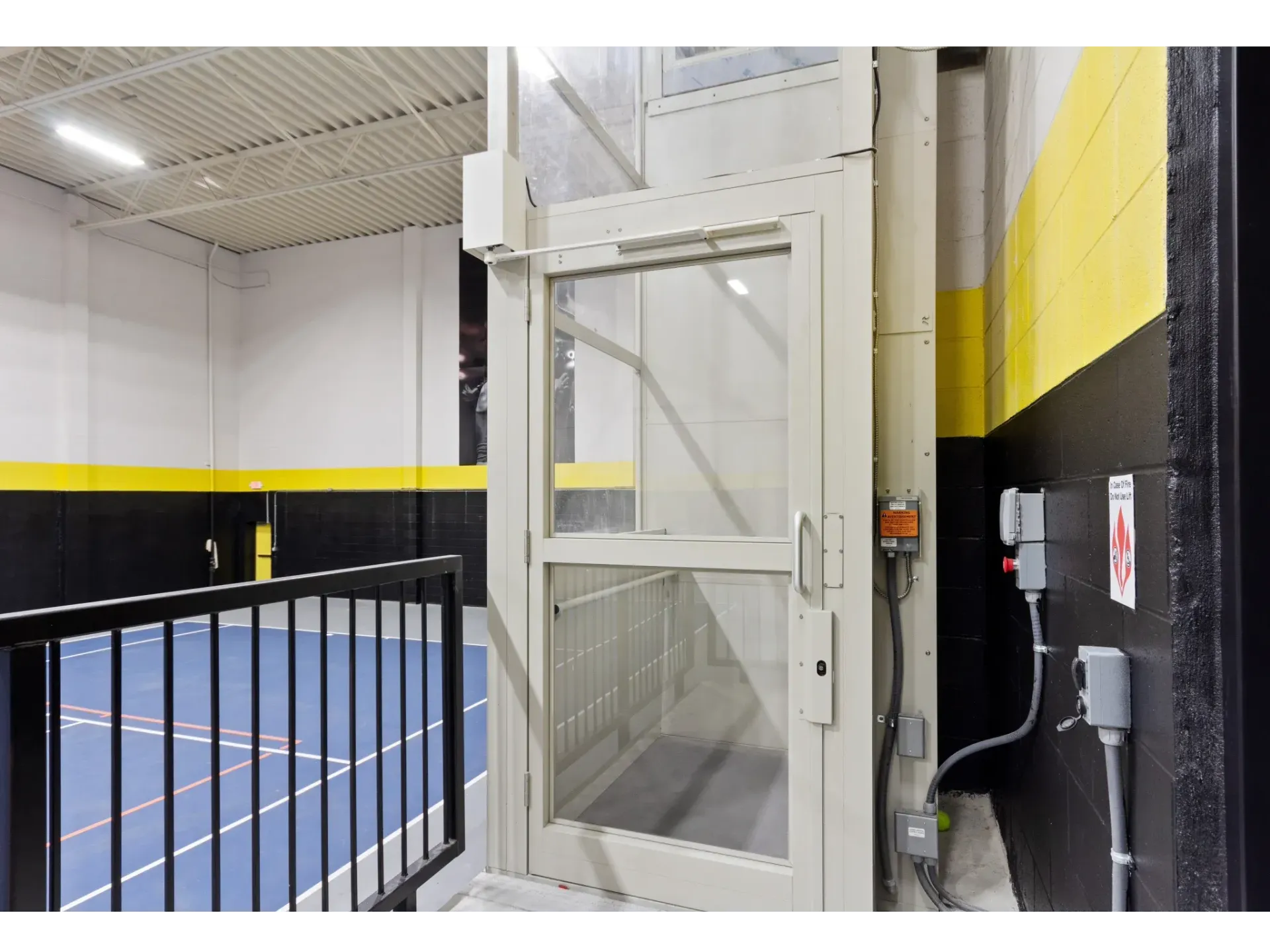 Indoor community recreation area with a blue court floor, yellow/black walls, and a glass service elevator.