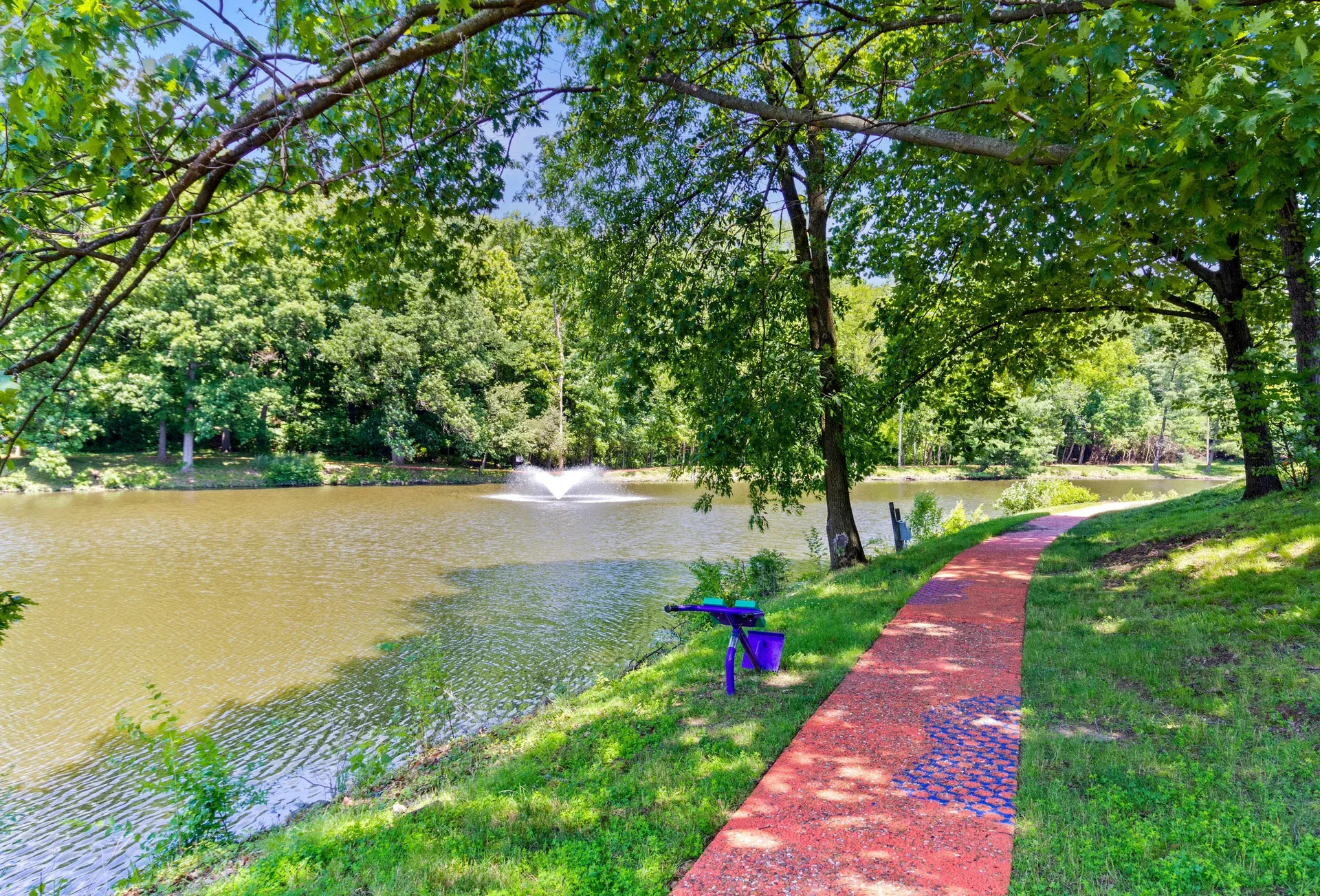 Pond with fountain, red walkway, and lush trees in a park-like apartment community.