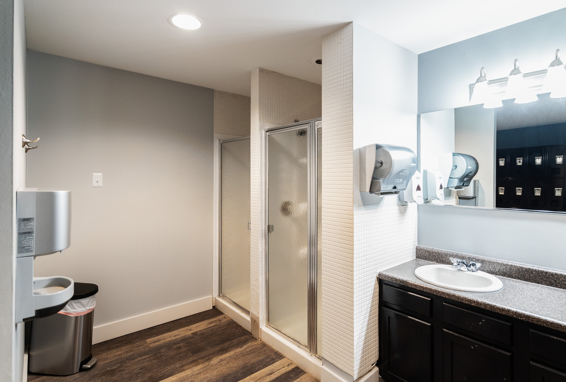 Public community locker room with a shower stall, sink, mirror, and wall-mounted dispensers.