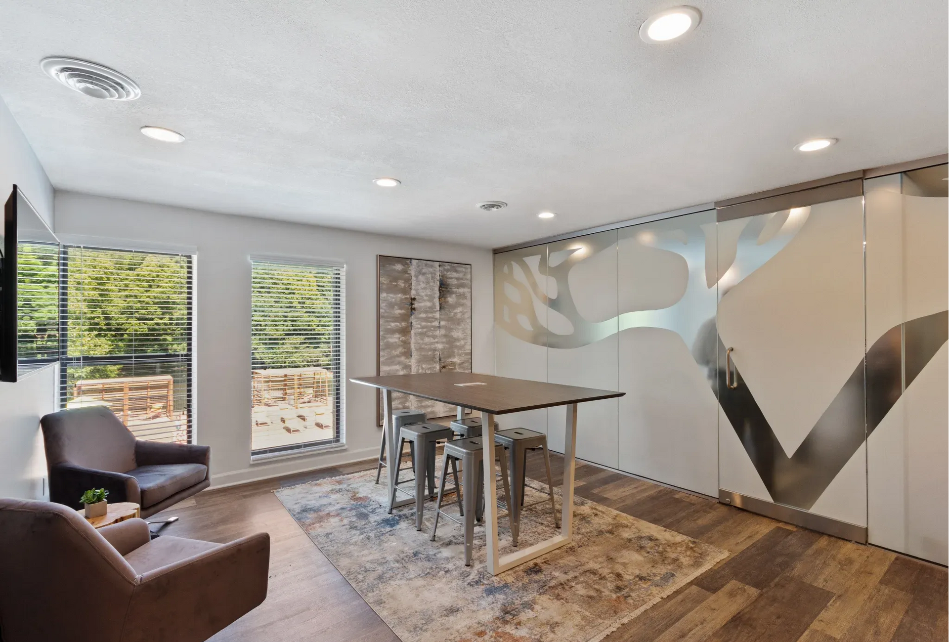 Community lounge with a high-top table, metal stools, two armchairs, large windows, and frosted glass wall.
