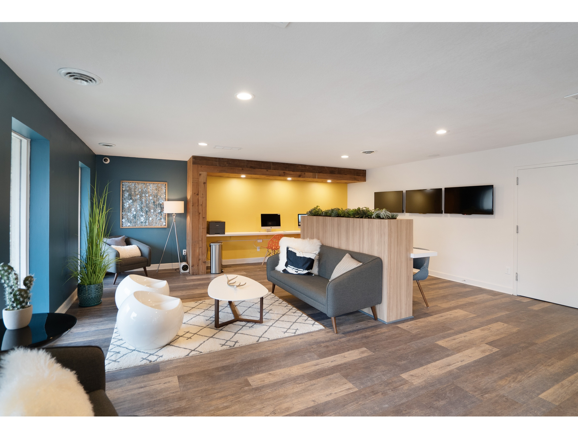 Modern apartment community lounge with seating, plants, and wall-mounted monitors.