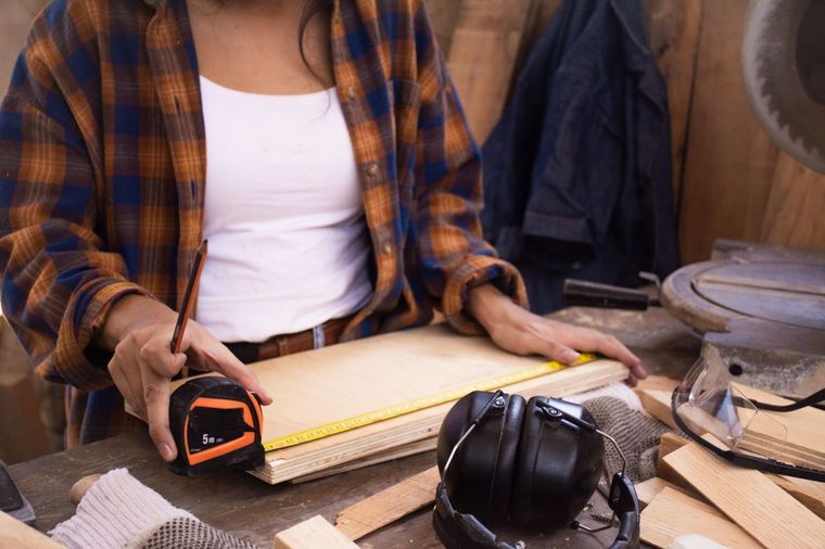 Person measuring a wooden board with a tape measure in a workshop, surrounded by tools and materials