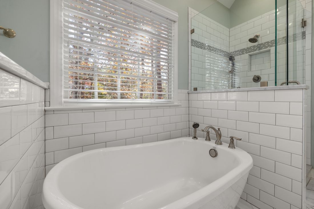White tiled bathroom with a freestanding tub, glass shower, and window blinds