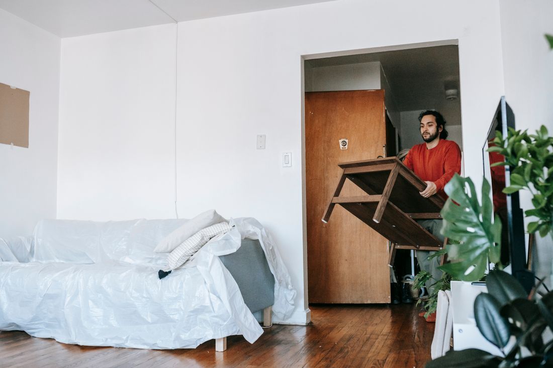 Person carrying a table through a doorway into a bright room with a white bed and wooden floor