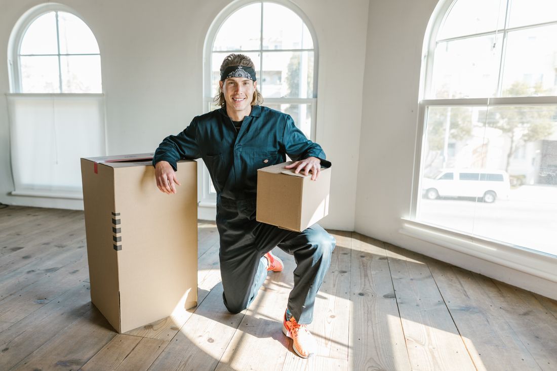 Person kneeling beside moving boxes in a bright empty room with arched windows.