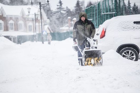 Person using a snowblower to clear a snow-covered driveway beside a white SUV