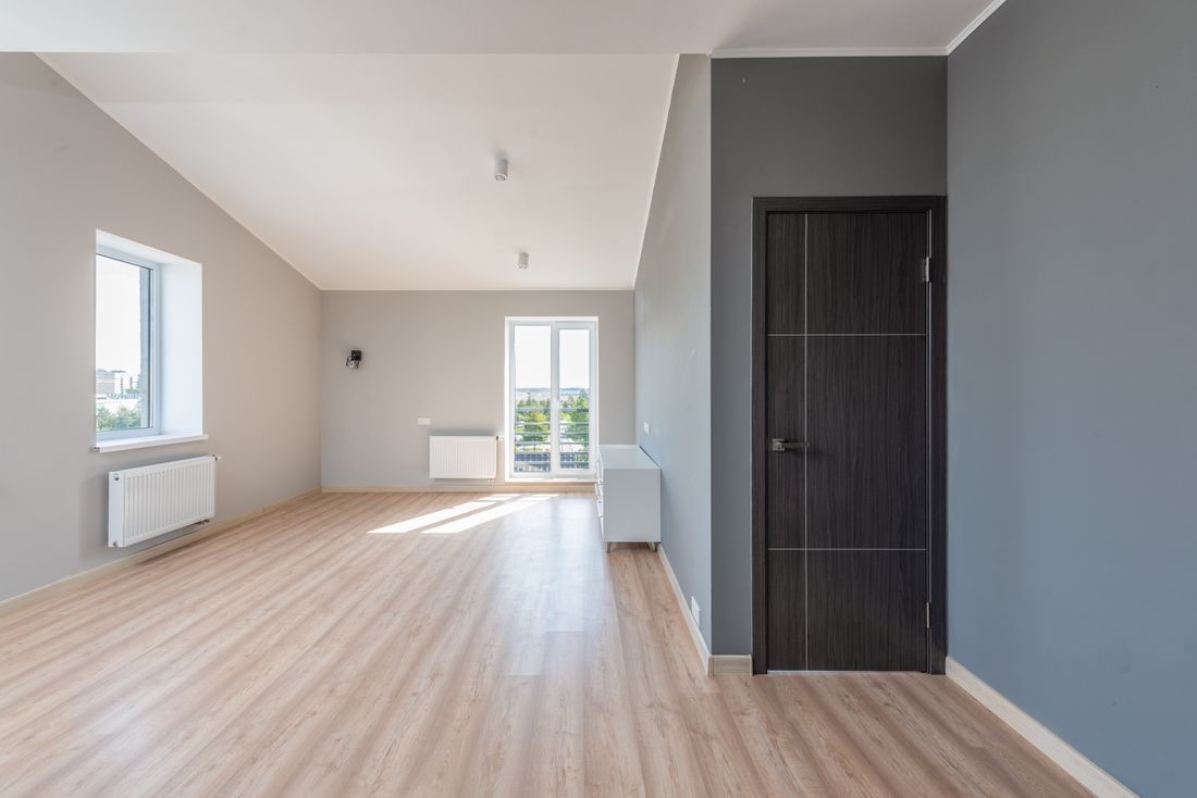 Empty bright room with light wood floors, white walls, windows, and a dark door.
