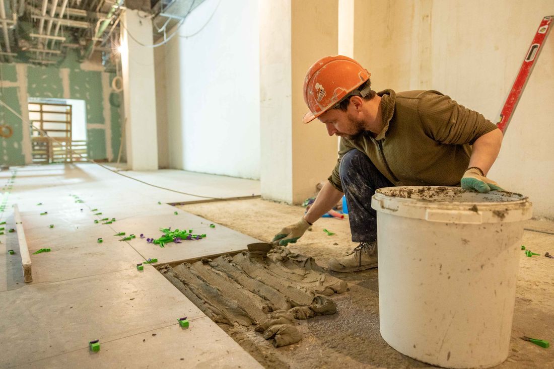 Worker smoothing concrete on a floor with a trowel beside a white bucket in an unfinished room