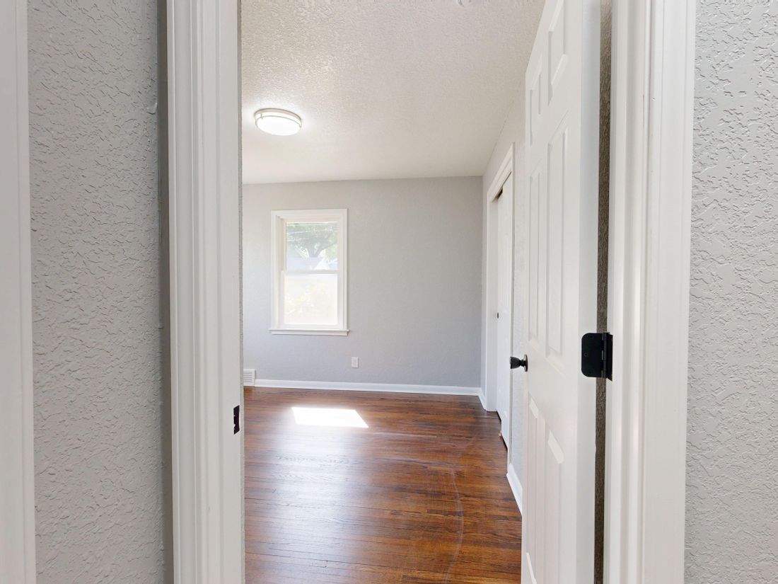 Empty hallway with white walls, wood floor, and a window in the back room.