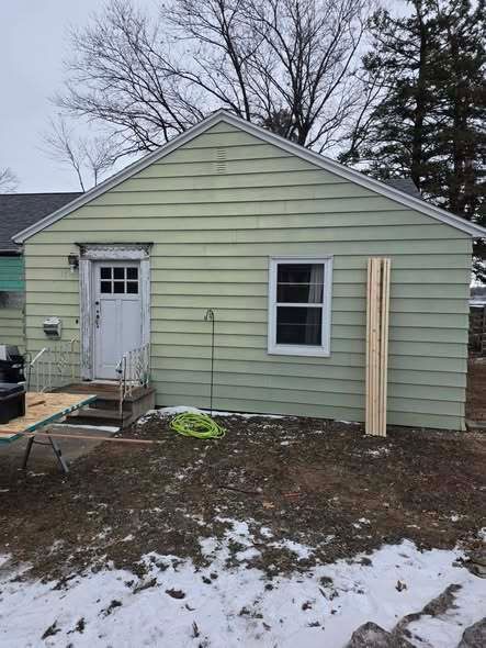 Small pale green house exterior with a white door, window, and snow-covered yard.
