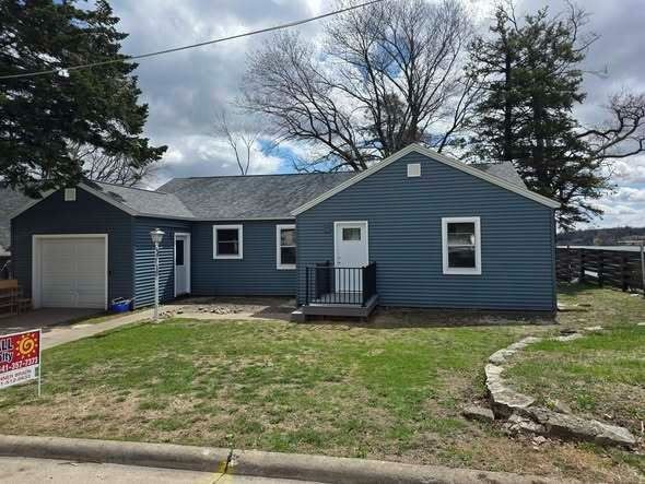 Blue single-story house with attached garage, front porch, and a patchy lawn under cloudy skies
