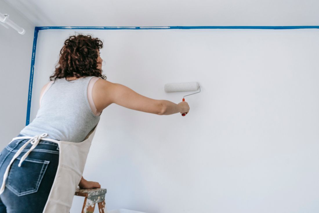 Person painting a white wall with a roller, masking off the top edge with blue painter’s tape.