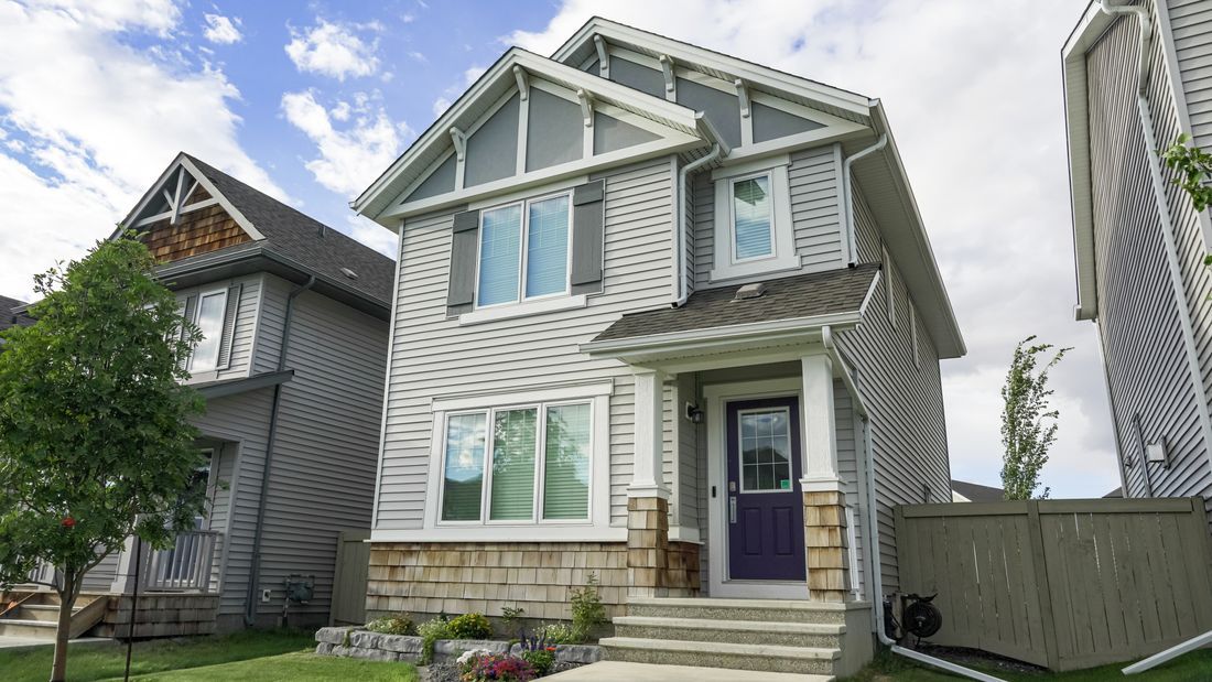 Two-story gray house with white trim, purple front door, and a small front yard under a blue sky