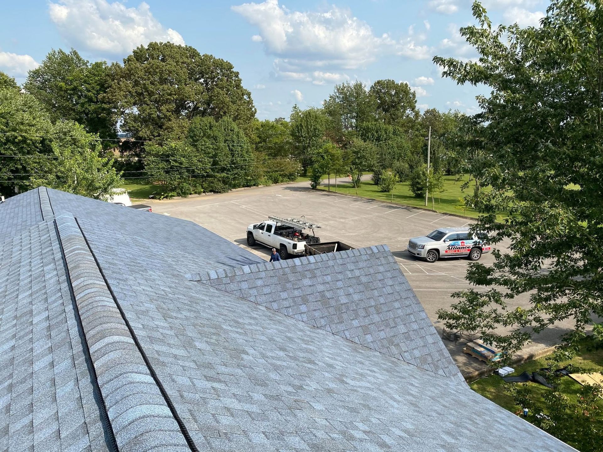 Roof shingles in the foreground overlooking a parking lot with a few cars and trees under a partly cloudy sky