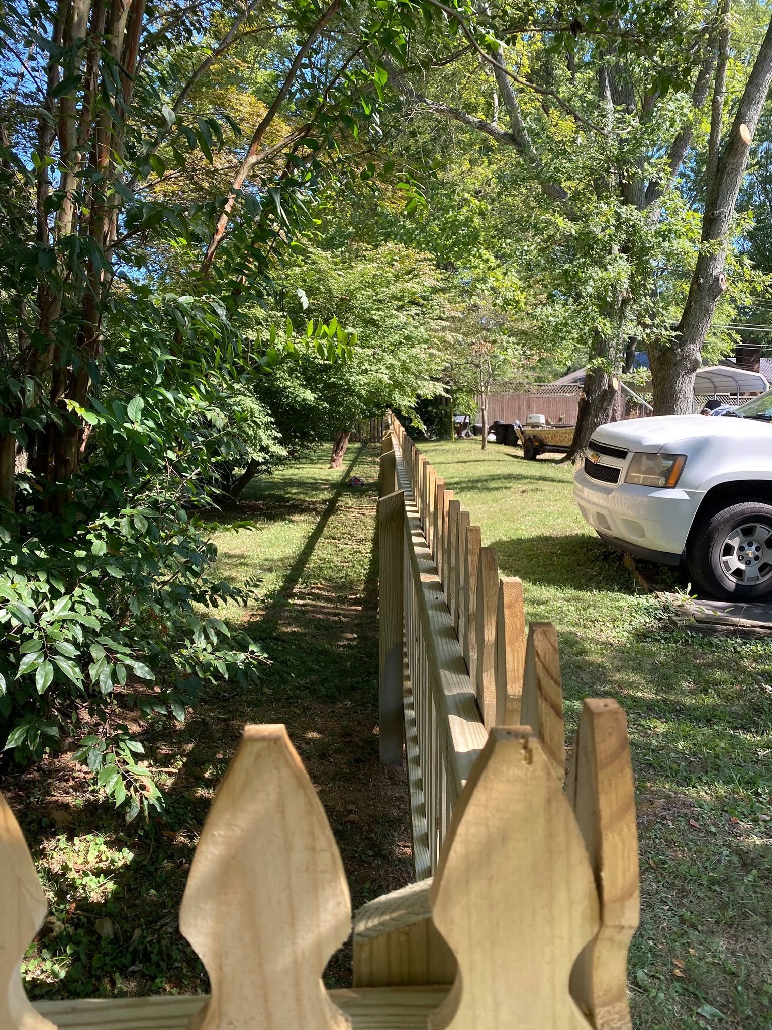 Wooden fence beside a shaded yard with trees, greenery, and a white truck parked on the grass.
