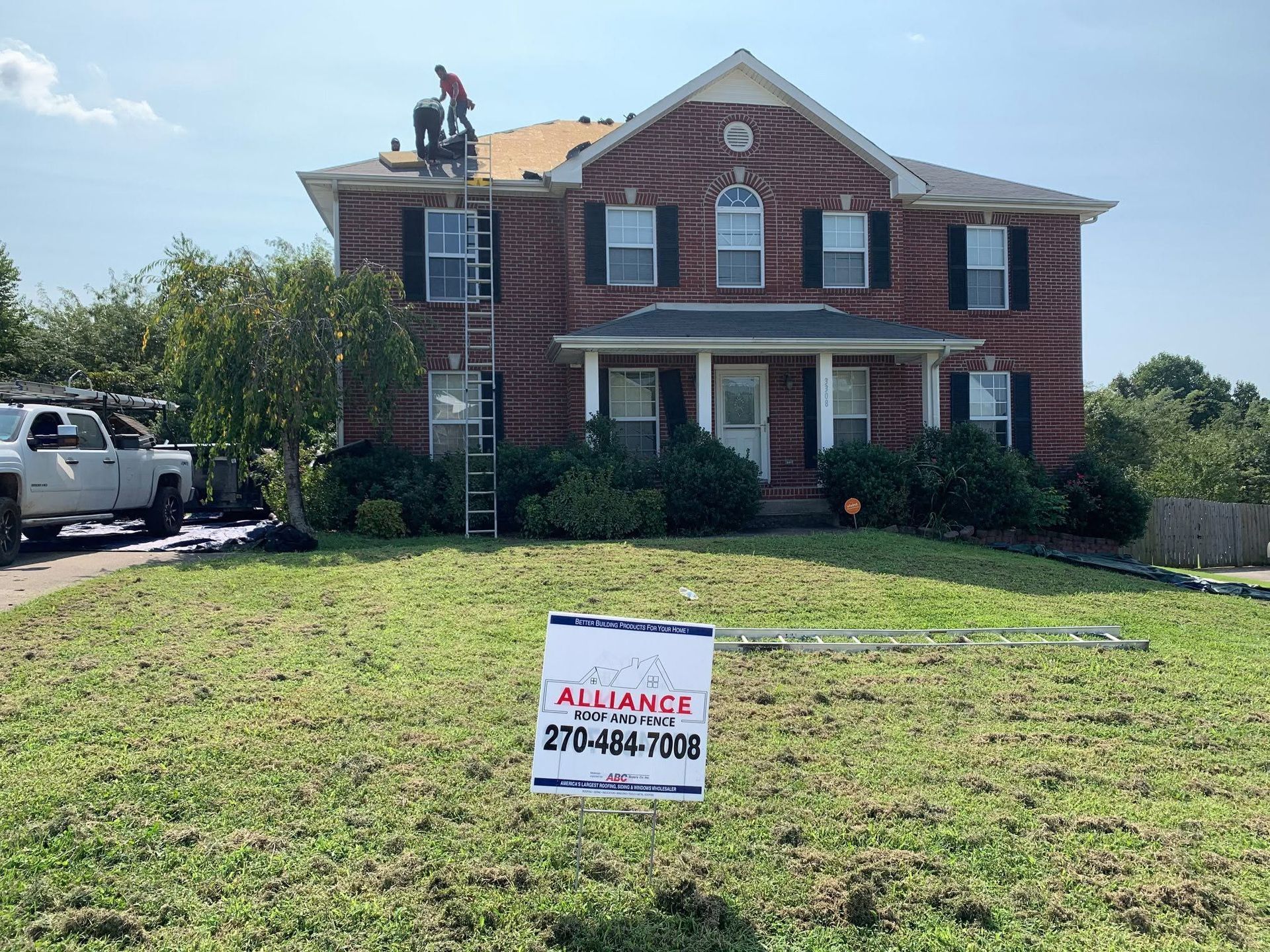 Two-story brick house with workers on the roof and a real estate sign in the front yard