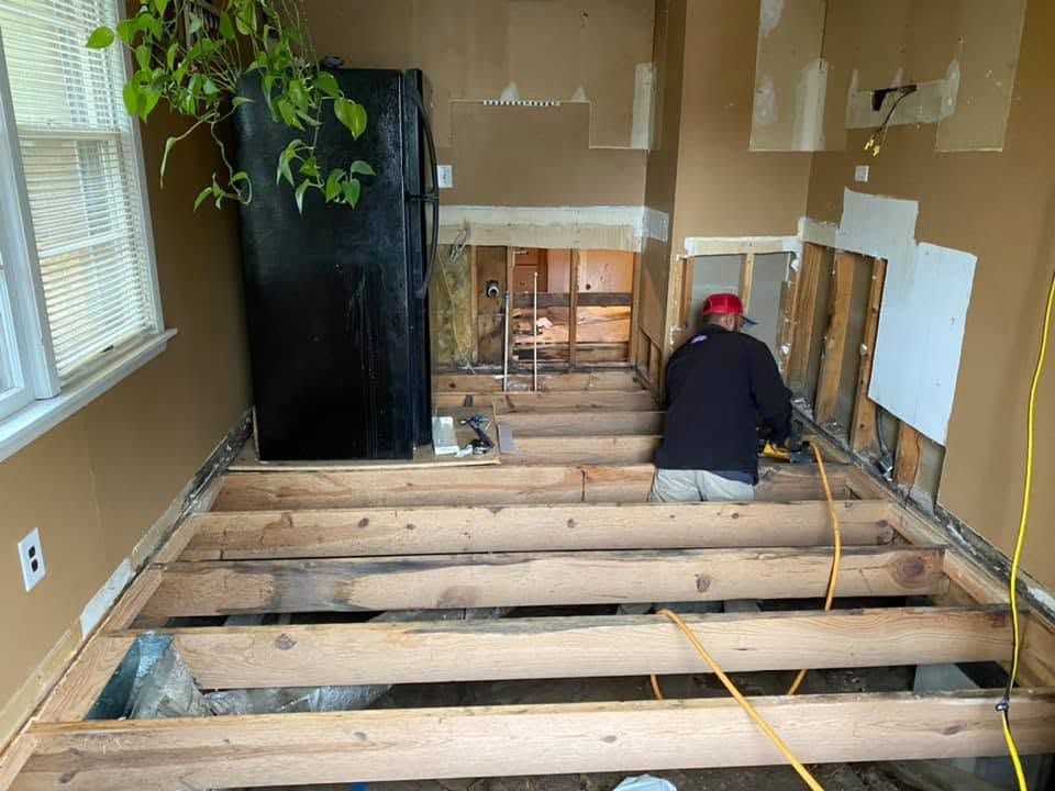 Demolished room with exposed floor joists; worker kneels near wall during renovation.