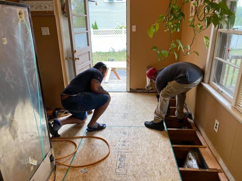 Two workers kneeling to repair a floor in a room with exposed joists and tools.