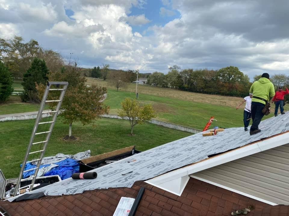 Workers repairing a roof with ladders and tarps beside a grassy field under cloudy skies