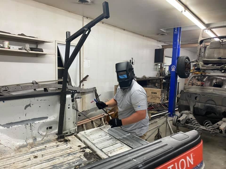 Person welding inside a white workshop beside a truck bed and car frame, wearing a welding helmet.