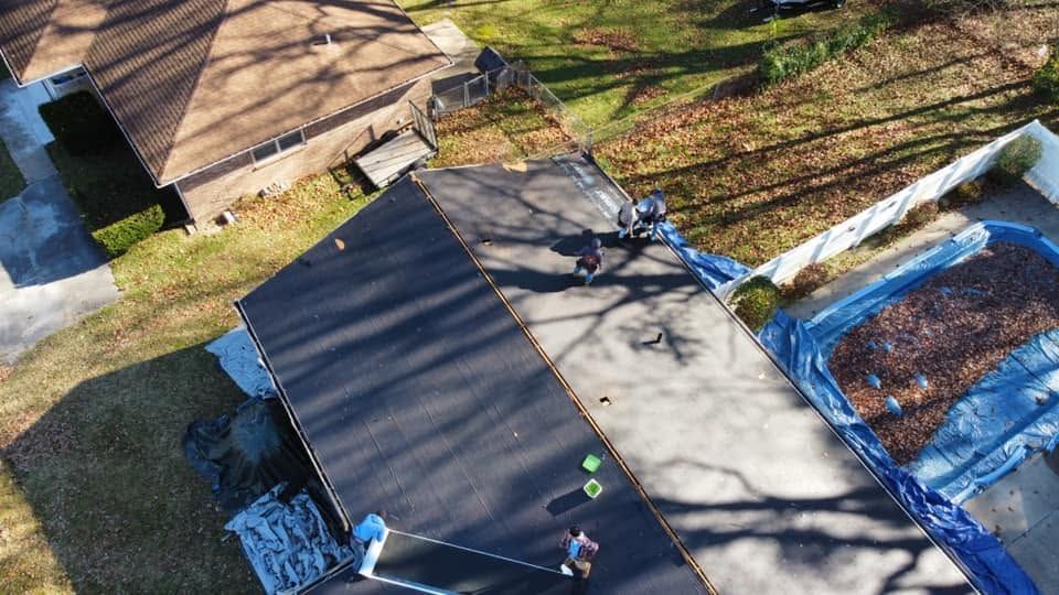 Aerial view of a roof installation with workers on a house roof beside a blue tarp-covered area.