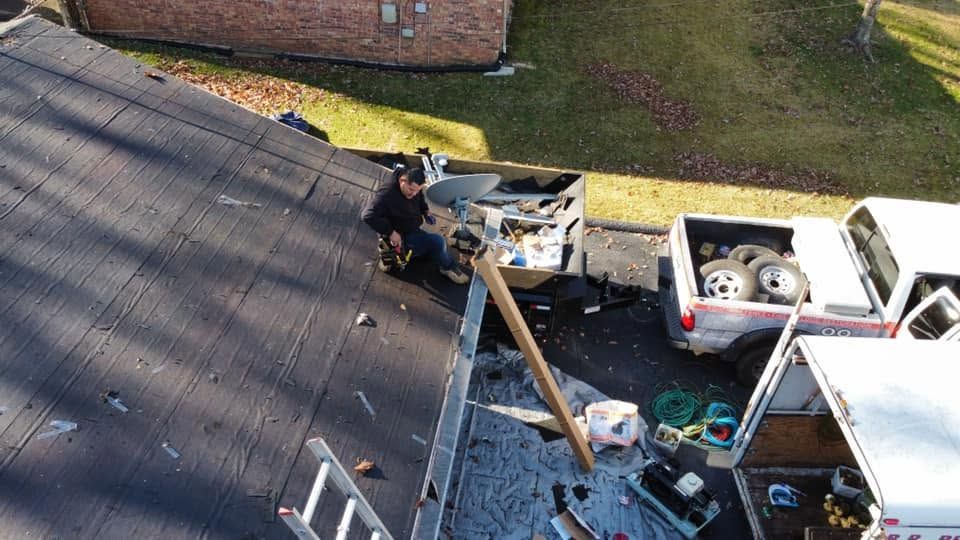 Worker on a shingled roof repairing an HVAC unit beside a white truck on a driveway